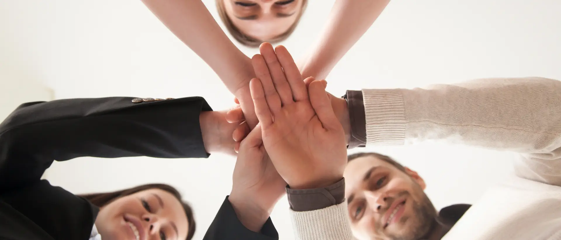 Vista en contrapicado de un equipo de cuatro profesionales sonrientes uniendo sus manos en el centro, simbolizando el trabajo en equipo y la colaboración empresarial.