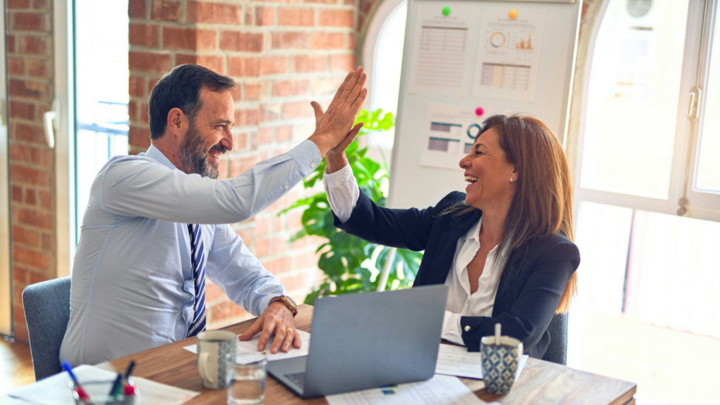 Consultores de Customer Excellence que representan la empatía, la resolución de problemas y la colaboración en la experiencia del cliente, celebran chocando sus manos en alto y con sonrisa genuina algún logro empresarial. La mesa donde ellos se encuentran sentados reflejan la aplicación práctica de nuestros talleres de formación en atención al cliente y fidelización. Fondo de oficina moderna.