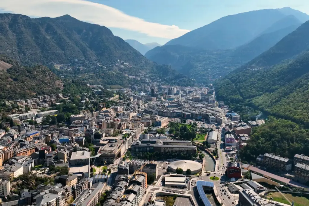 Vista aérea panorámica de Andorra la Vella situada en un valle profundo de los Pirineos, mostrando la ciudad densa con edificios modernos rodeados de montañas boscosas escarpadas bajo un cielo azul parcial