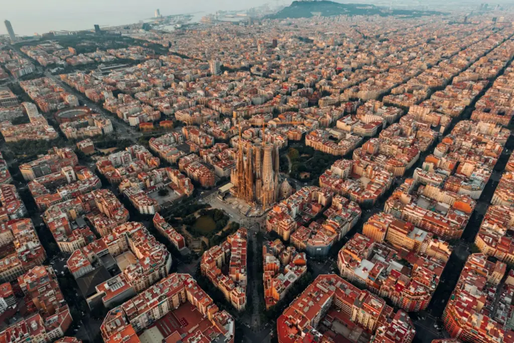 Vista aérea panorámica de Barcelona, mostrando la cuadrícula ortogonal del distrito del Eixample y la Sagrada Familia de Gaudí iluminada en el centro al atardecer, con el mar Mediterráneo y la montaña de Montjuïc al fondo