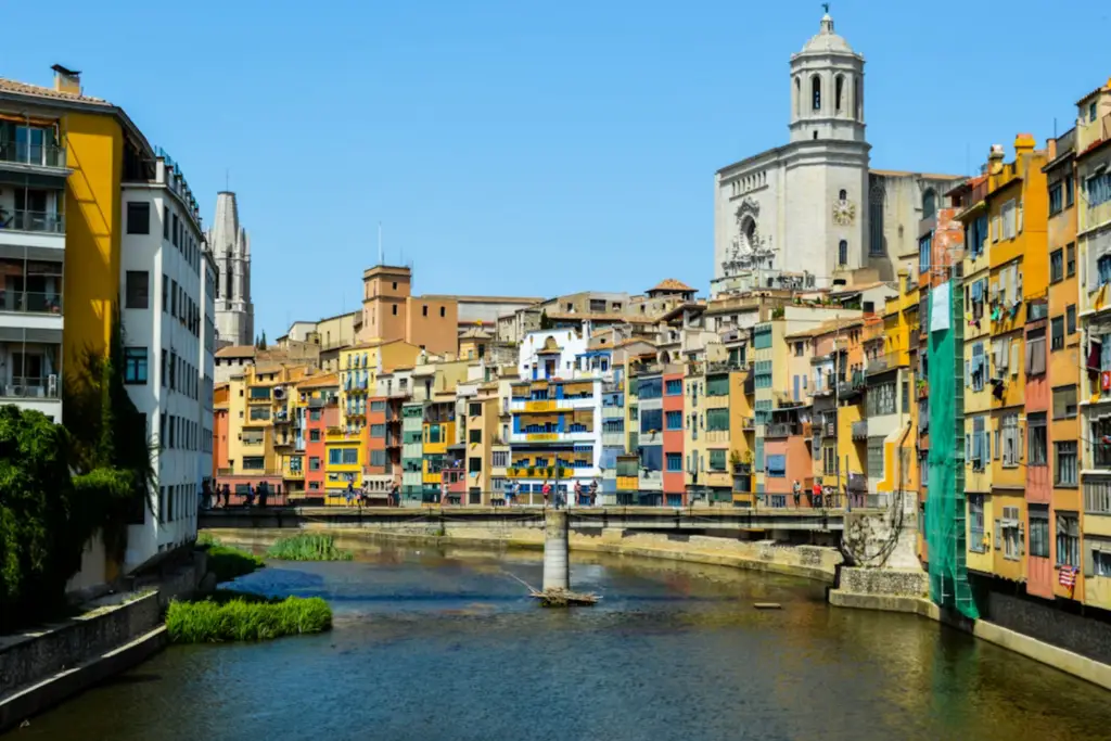 Vista de las coloridas casas del río Onyar en Girona, con el puente de piedra y la torre de la Catedral de Santa María al fondo bajo un cielo despejado.