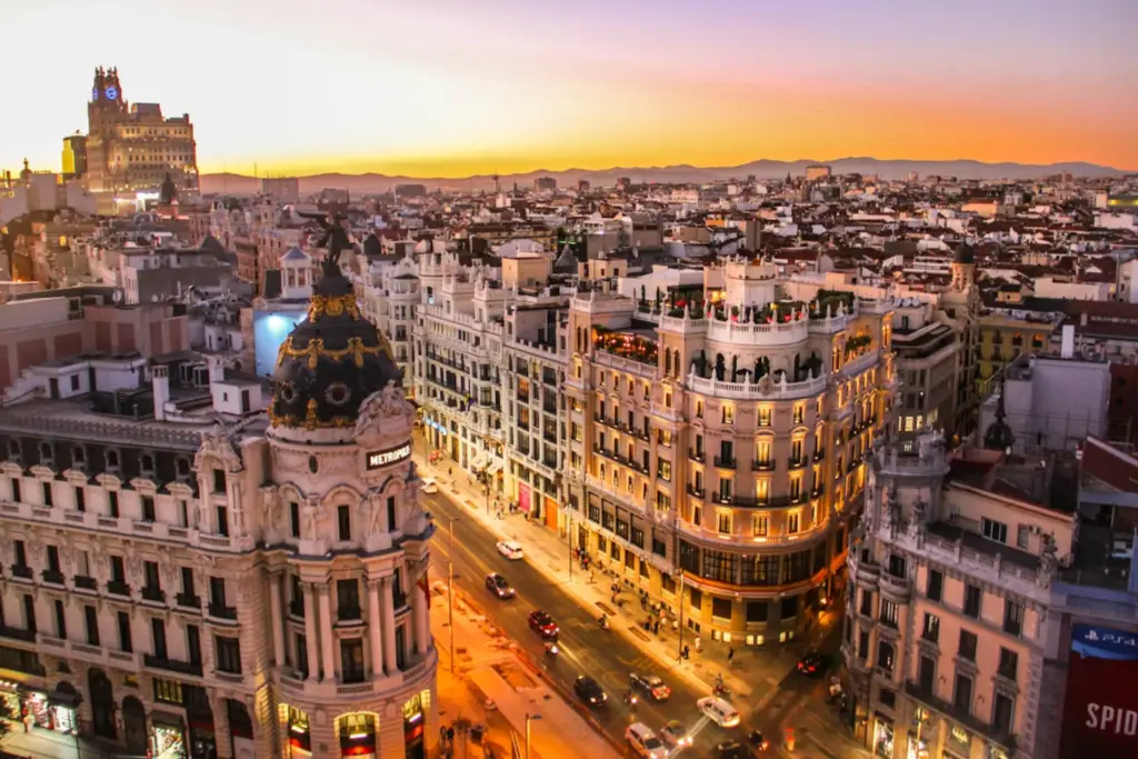 Vista aérea de la Gran Vía de Madrid al atardecer, destacando el Edificio Metrópolis con su cúpula negra y dorada en primer plano, bajo un cielo anaranjado y edificios históricos iluminados